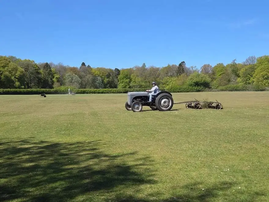 The club's vintage grey tractor towing gang mowers across the outfield under a blue sky
